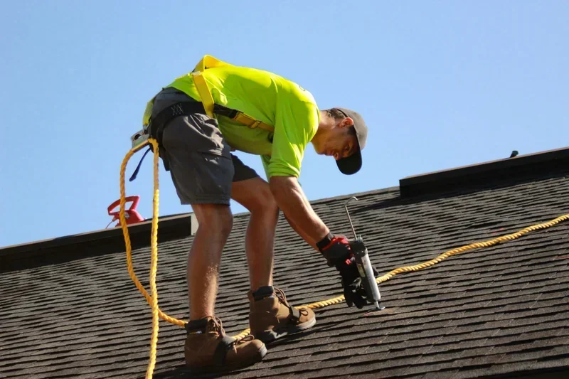 Roofer in a safety harness fastening asphalt shingles with a nail gun